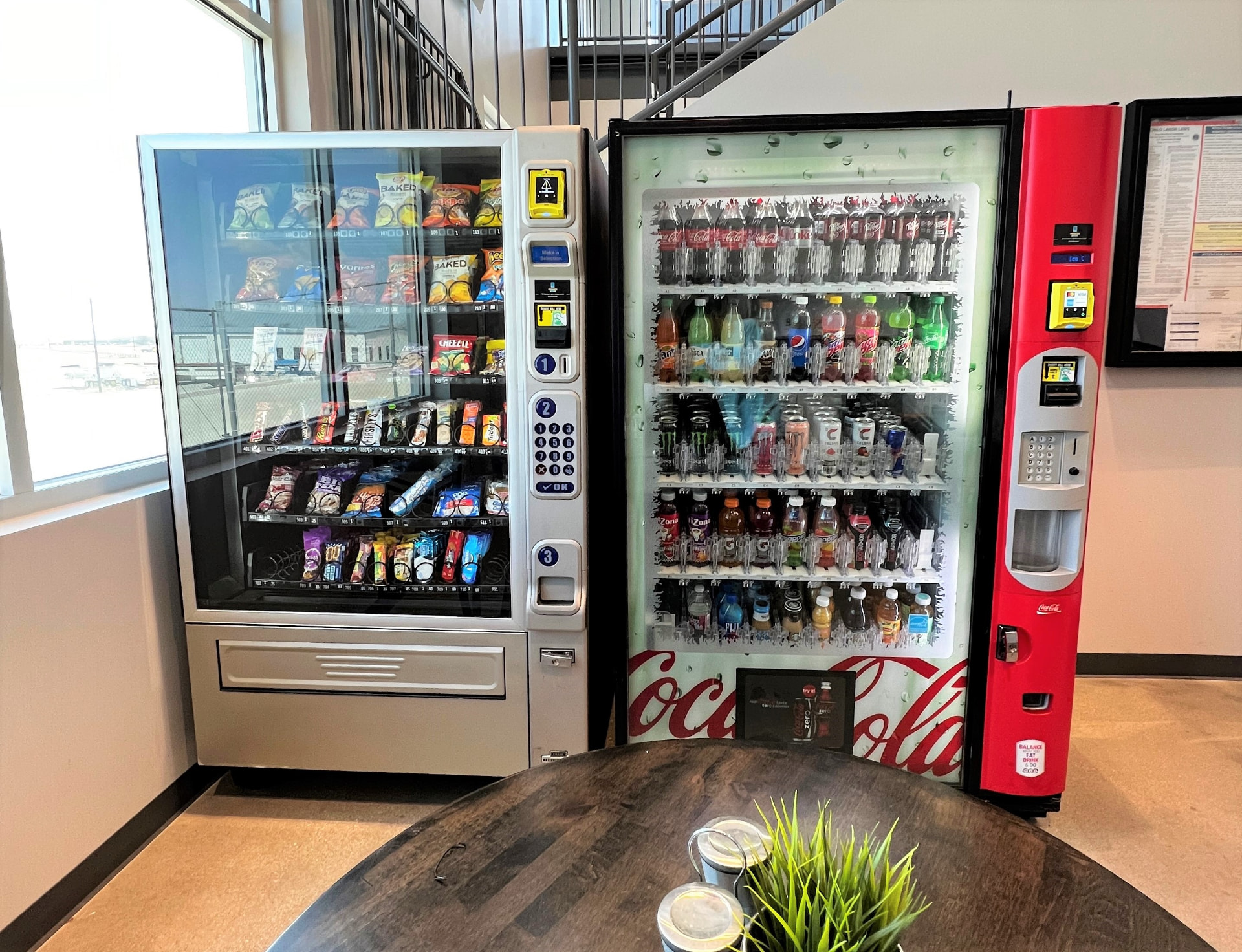A MunchBox AI smart cooler installed in a bright modern office breakroom, stocked with drinks, snacks, and fresh food